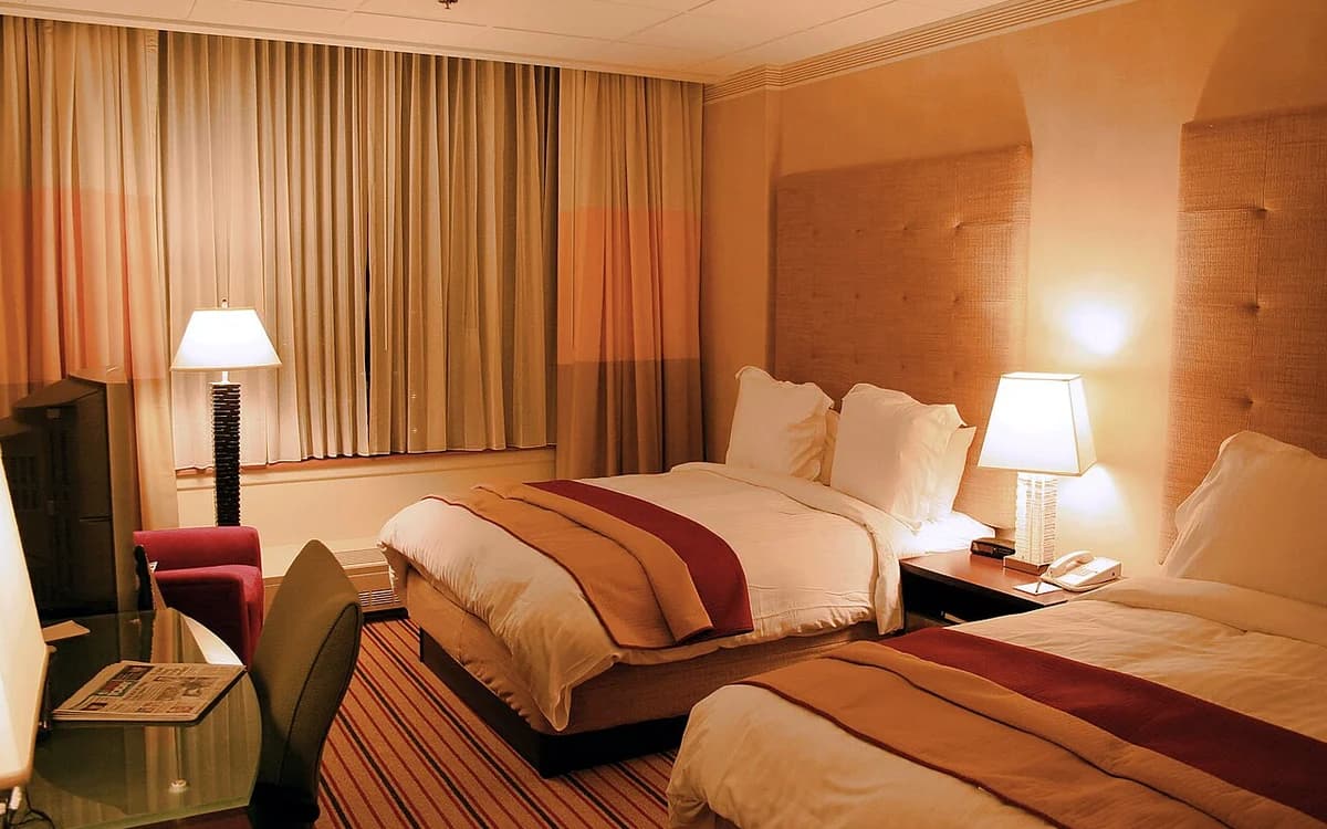 Modern hotel guestroom interior showing upholstered bed headboard, carpet, and full-height window blackout curtains
