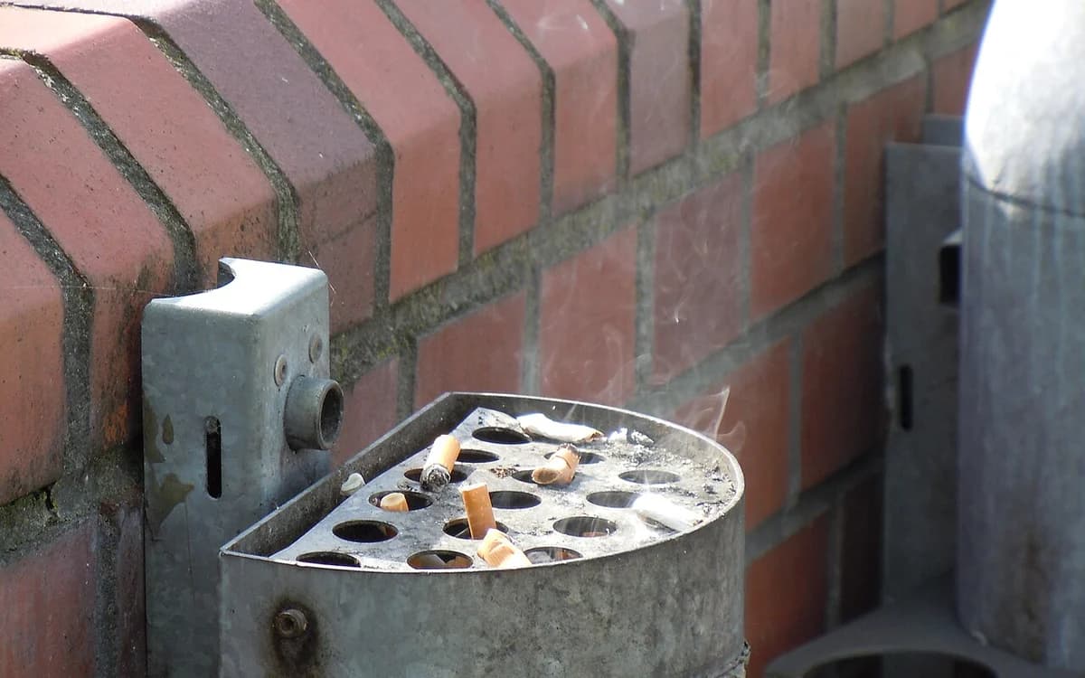 Smoldering cigarette in a metal ashtray showing side-stream tobacco smoke and tar residue